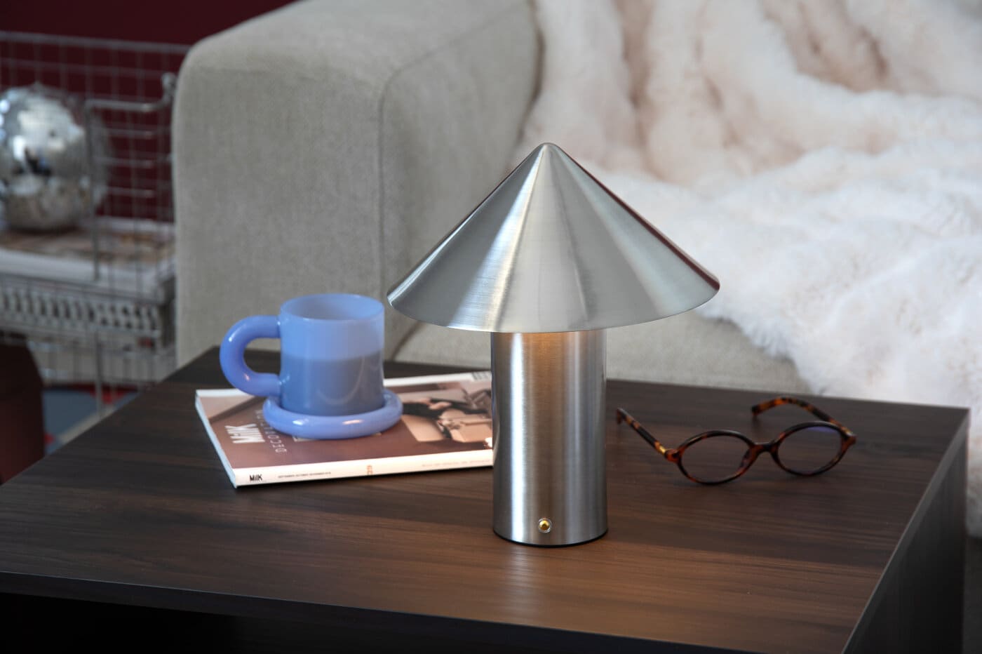 A portable metal table lamp beside a blue mug, a magazine and a pair of glasses on a wooden table.