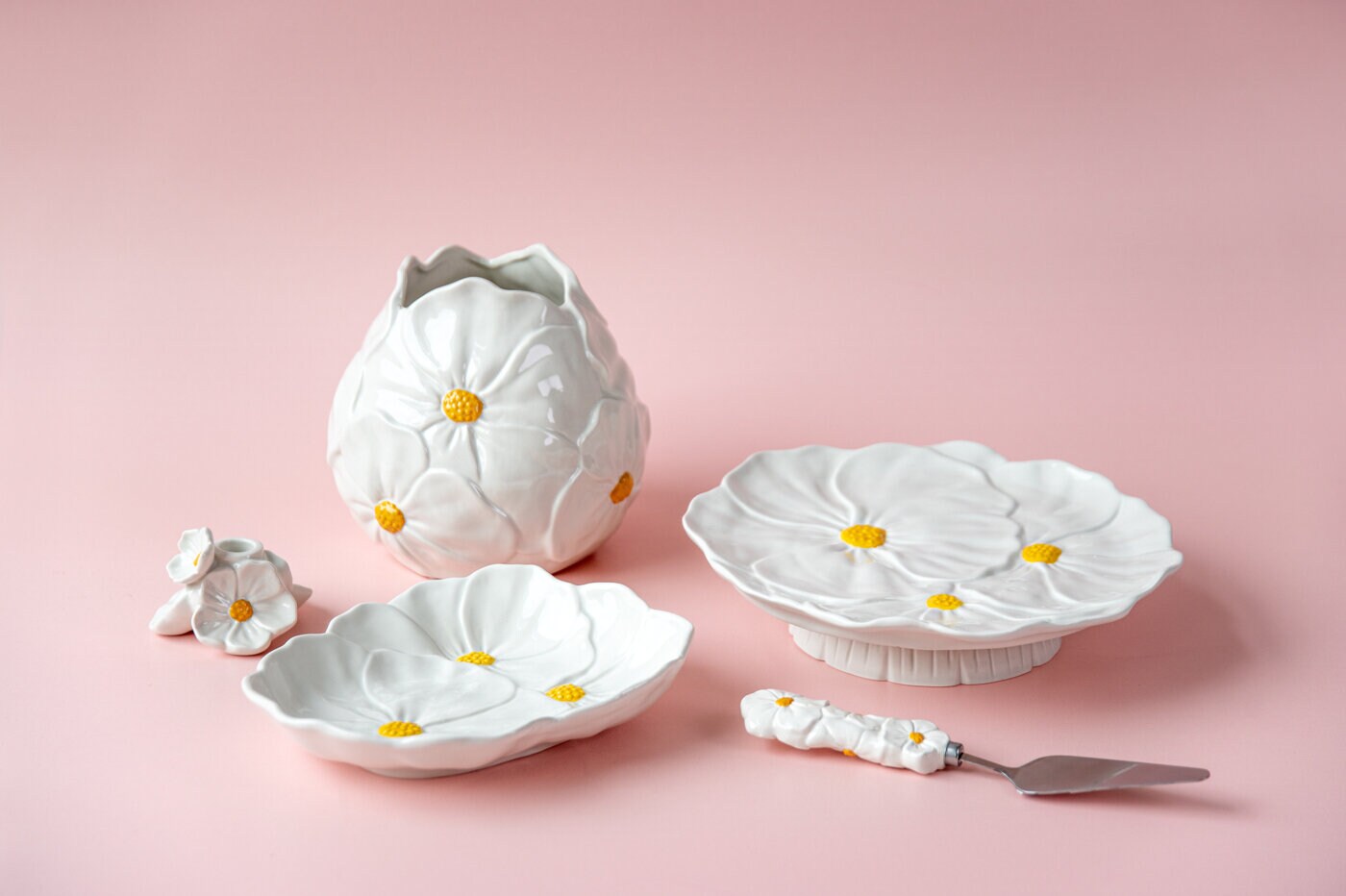 White ceramic vase and serving platter with flower-shaped details and yellow centers from the Blomstra series against a light pink background.