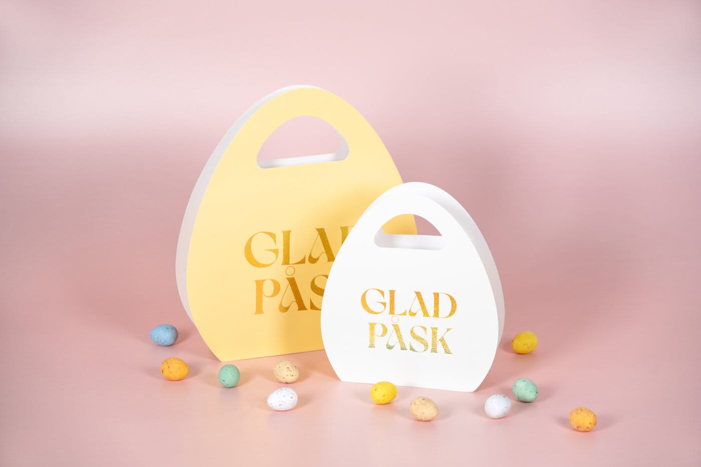 Egg-shaped gift bags with the text "Glad påsk", surrounded by small Easter candy on a soft pink background.