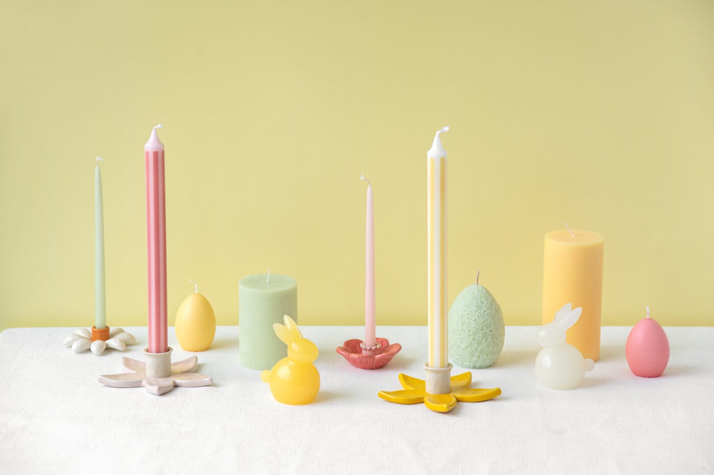 Pastel-coloured candles in various shapes, including egg- and bunny-shaped candles with decorative candle holders on a light table against a yellow background.