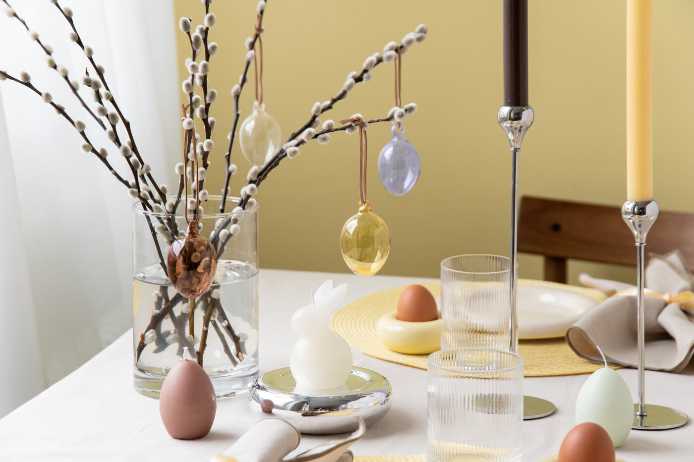 Easter table setting with willow branches, hanging Easter eggs, glass vase and egg on the table.