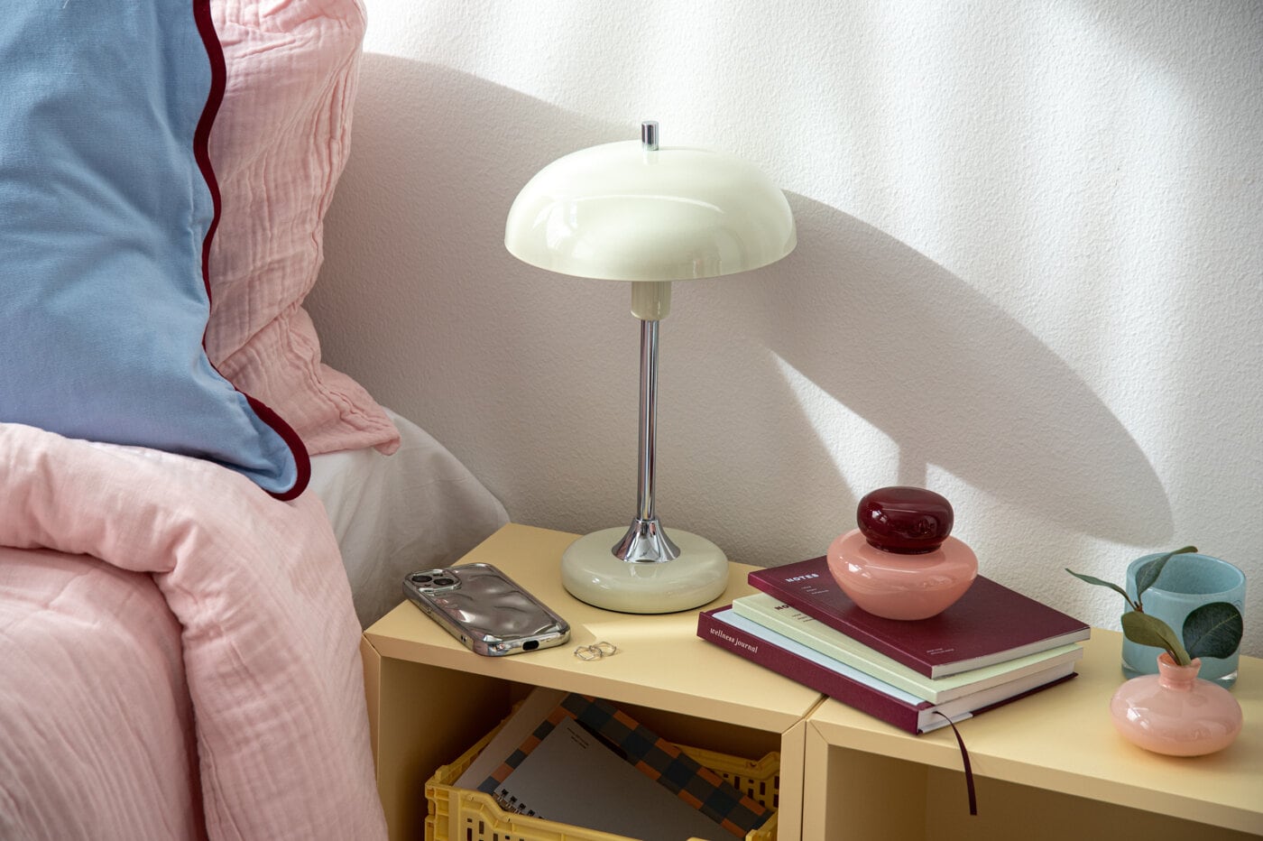 Bedside table with a table lamp, a mobile phone, and stacked books with a decorative jar on top.