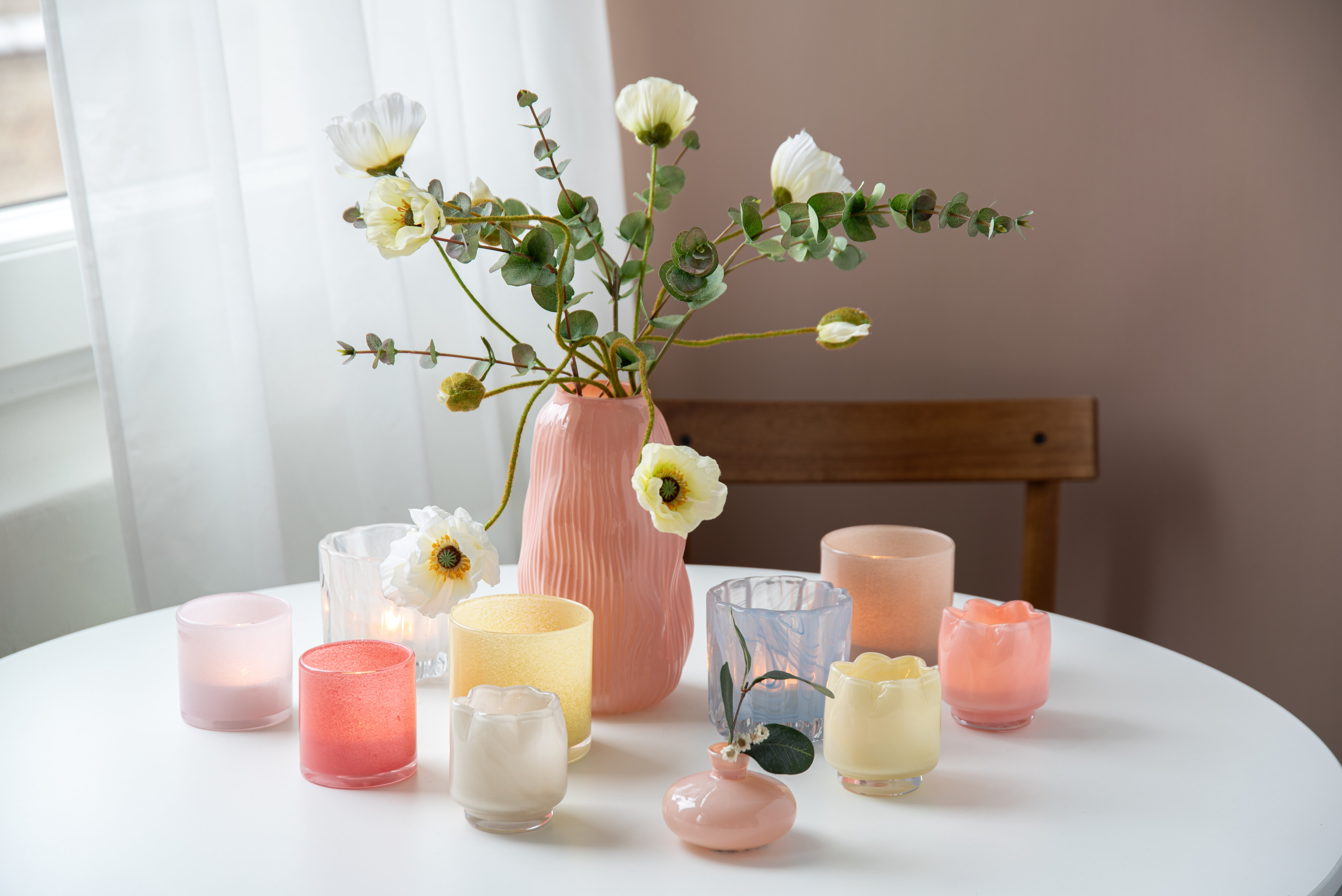 Pink vase with white flowers and green branches on a table, surrounded by glass candle holders in pastel colors.