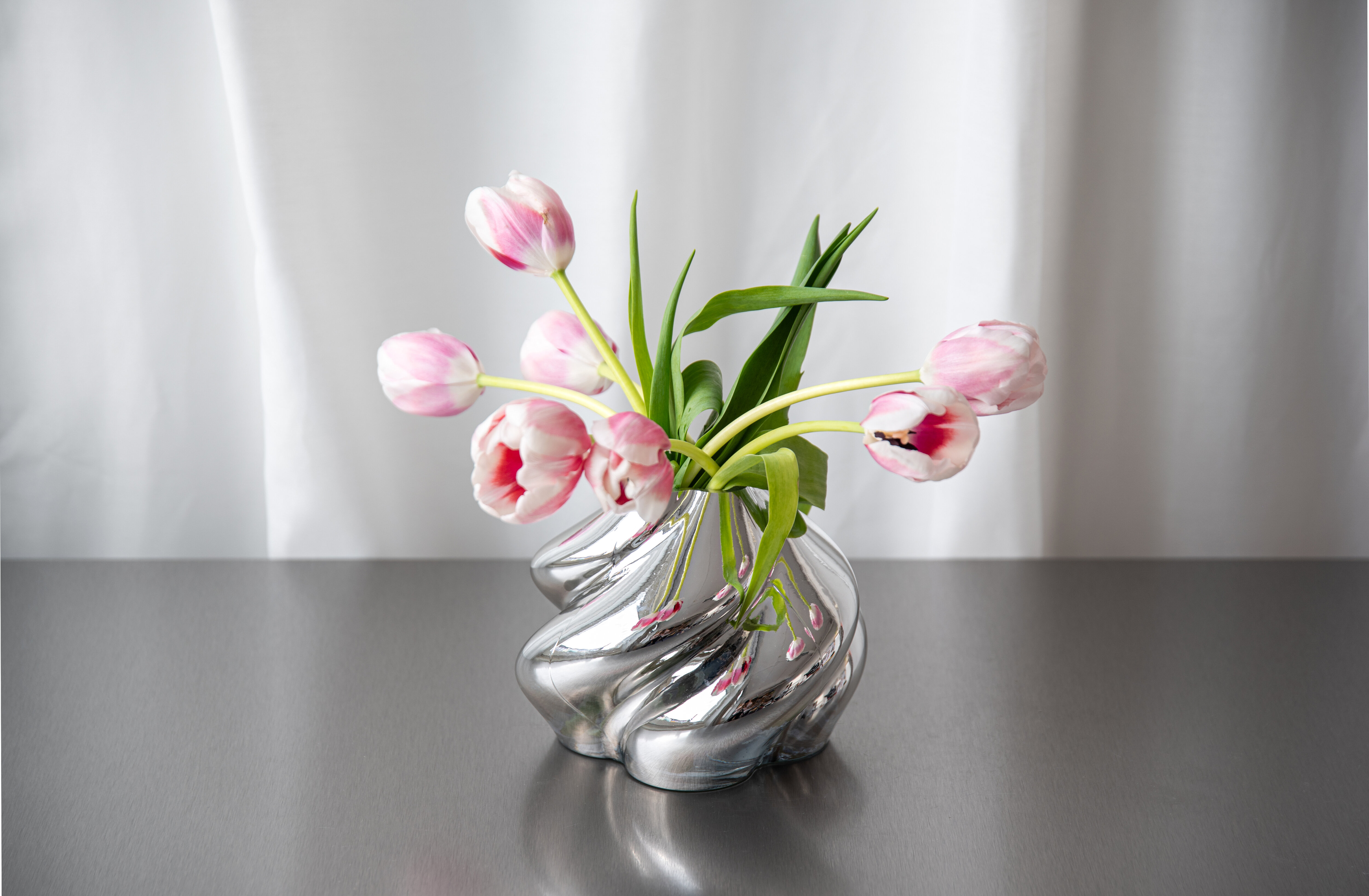 Pink tulips in a shiny, sculptural silver vase on a table.