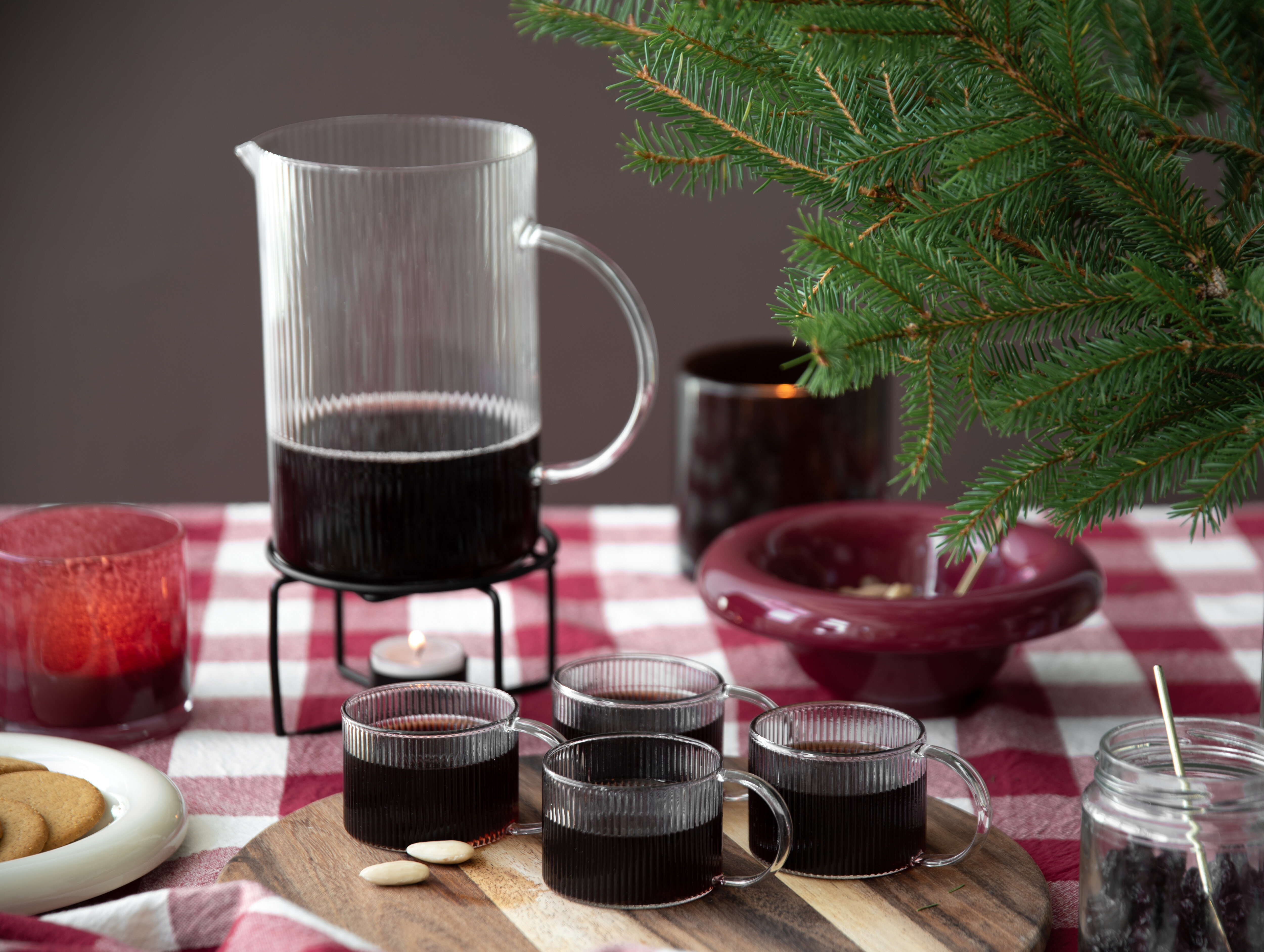 Mulled wine served in a glass jug and glass mugs on a festive table with fir branches.