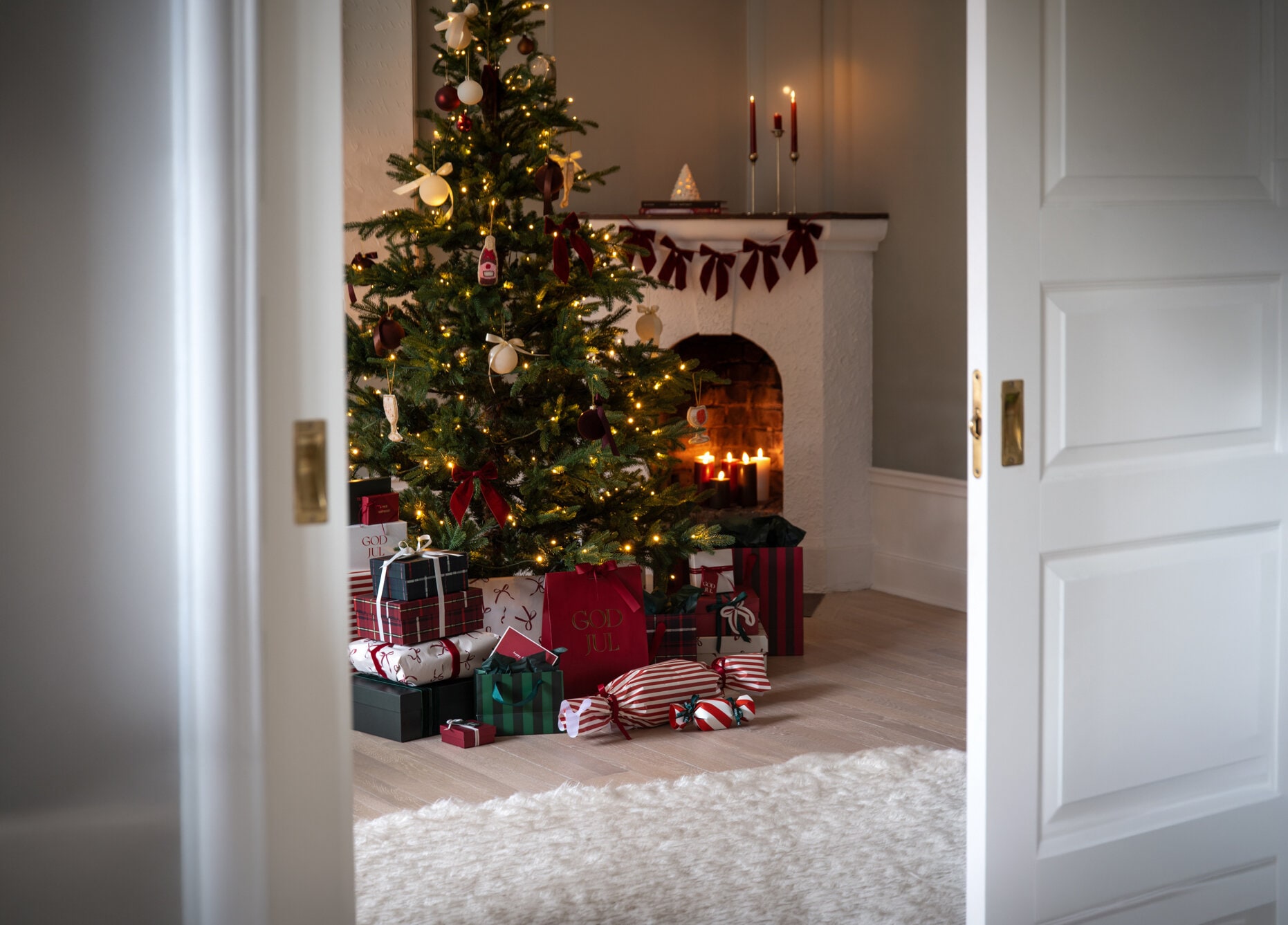 Decorated Christmas tree with string lights, red bows and wrapped gifts in front of a fireplace.