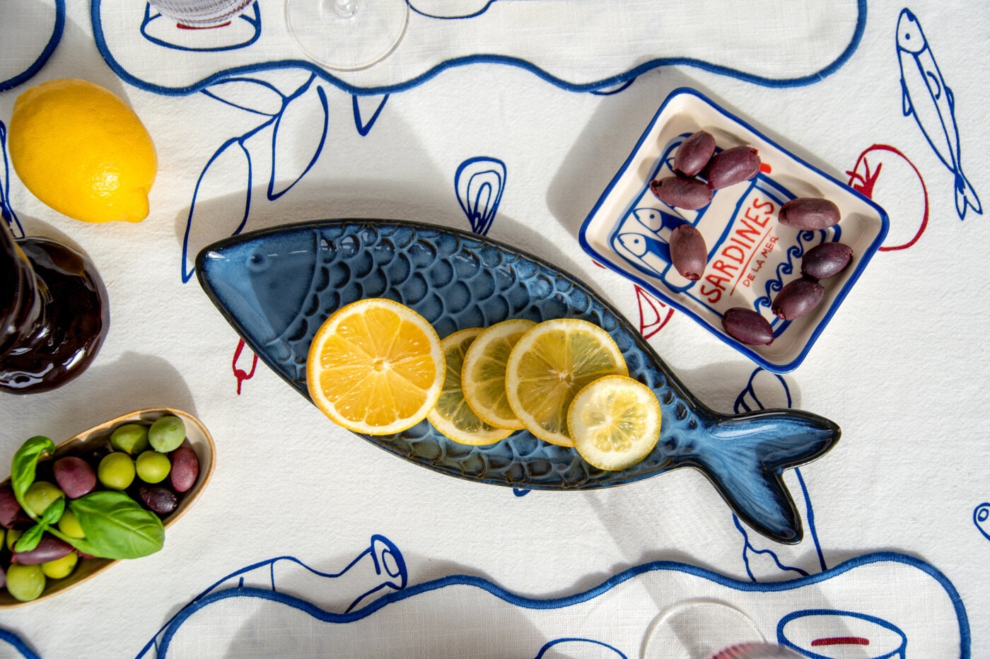 Fish-shaped serving platter with lemon slices on a patterned tablecloth, surrounded by olives and small bites.