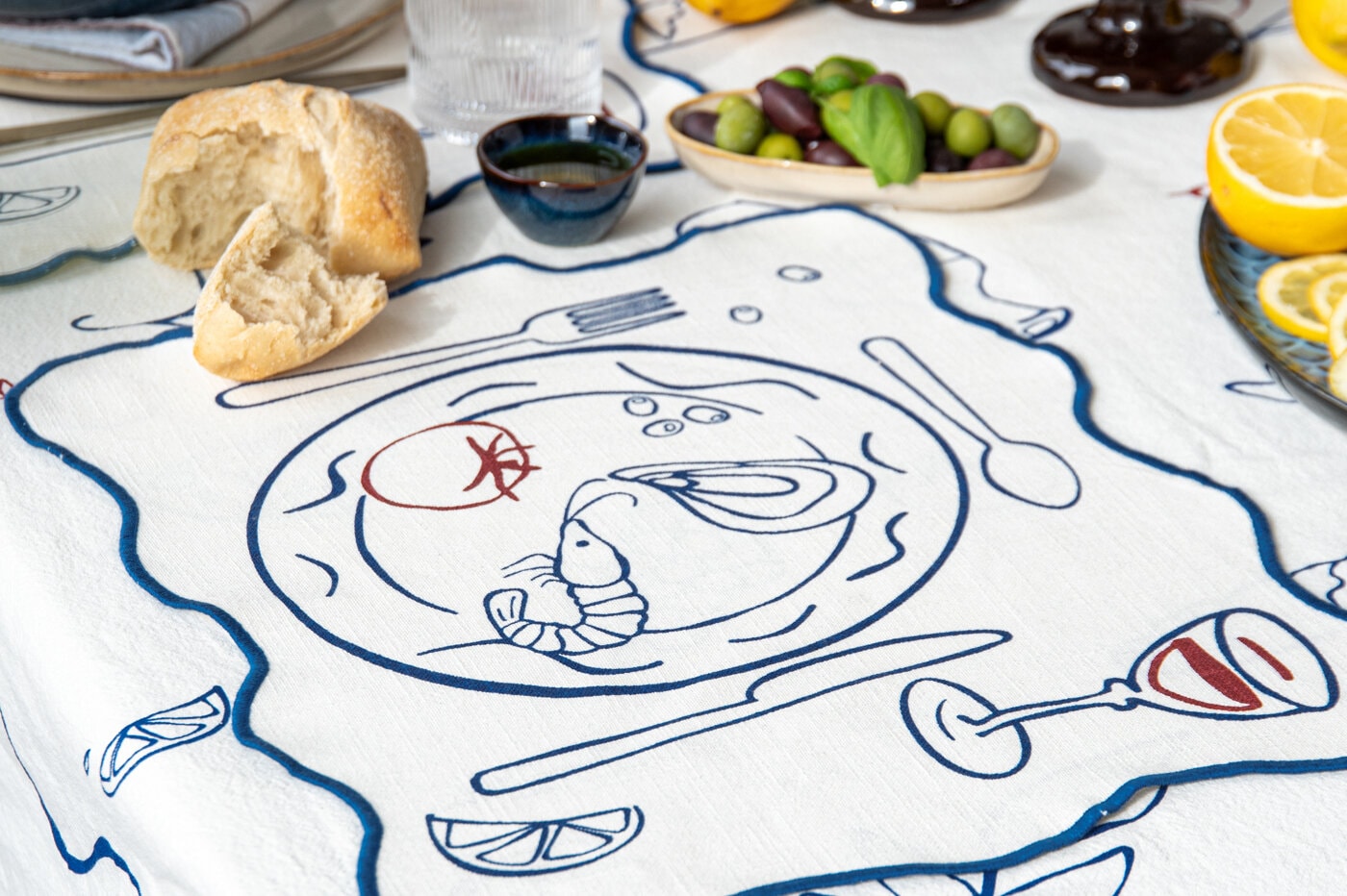 White tablecloth with blue and red illustrations of a table setting with a plate, cutlery, glass and food.