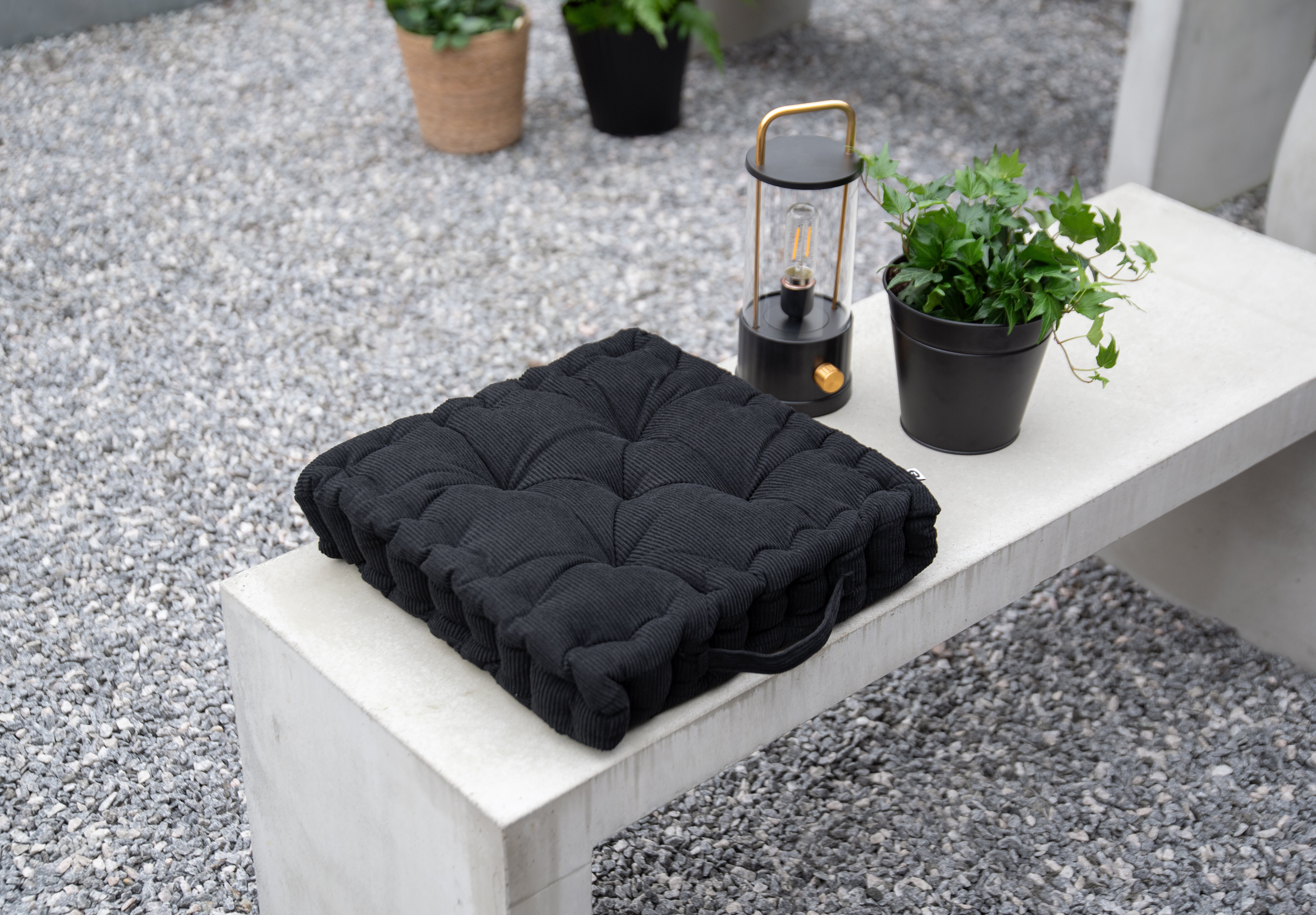 Black seat cushion on a concrete bench outdoors, with a decorative lantern and green potted plants.