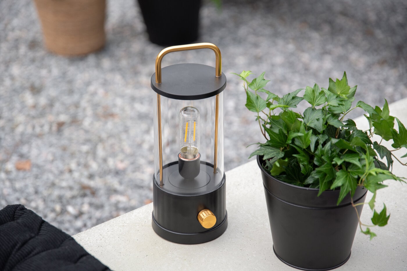 Lantern with handle and black pot with a green plant in an outdoor setting.