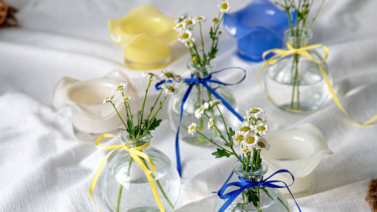 Små glasvaser med blommor och blågula band bredvid ljuslyktor i olika färger på ett dukat studentbord.