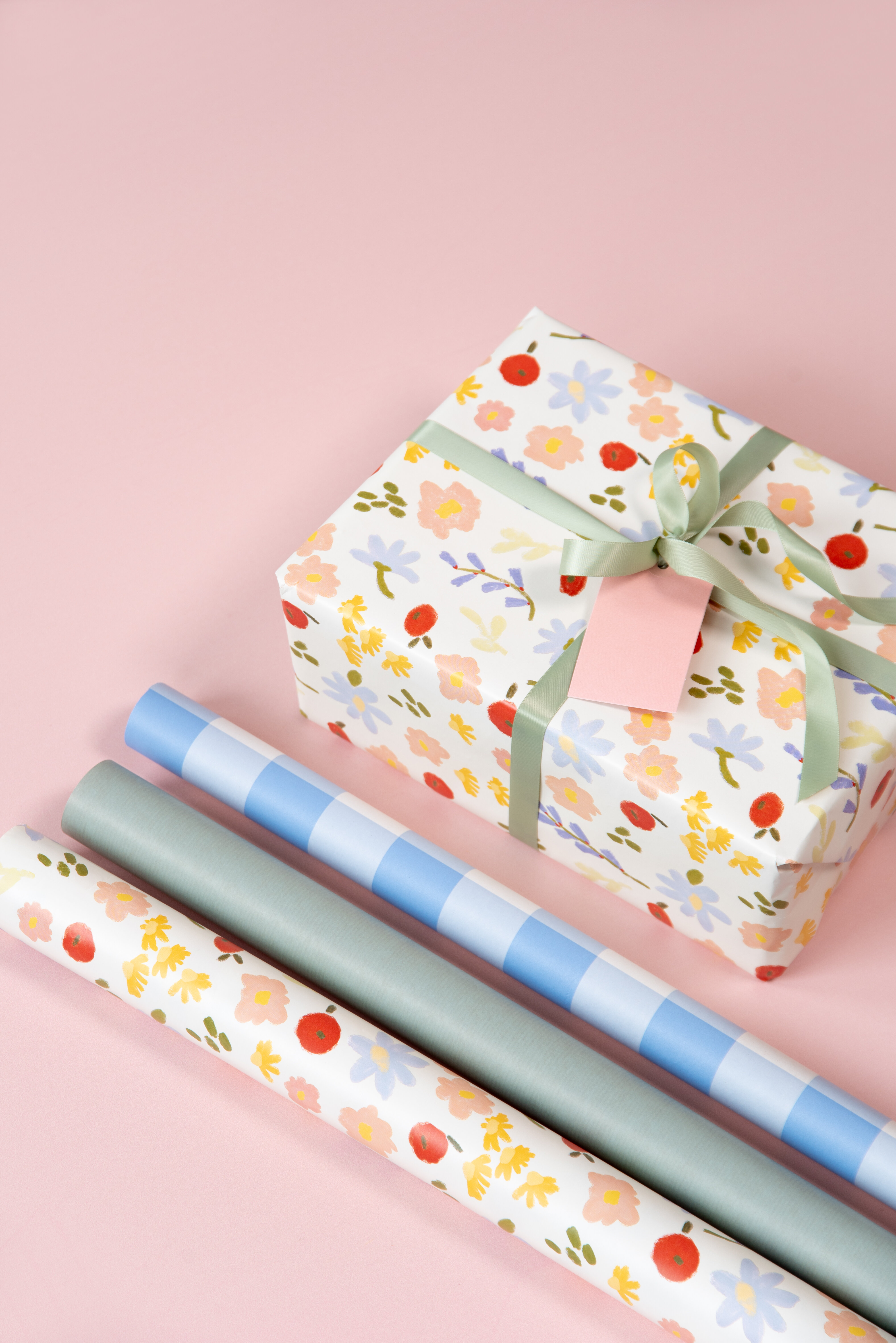 Gift box wrapped in floral paper with ribbon, alongside rolls of wrapping paper on a pink background