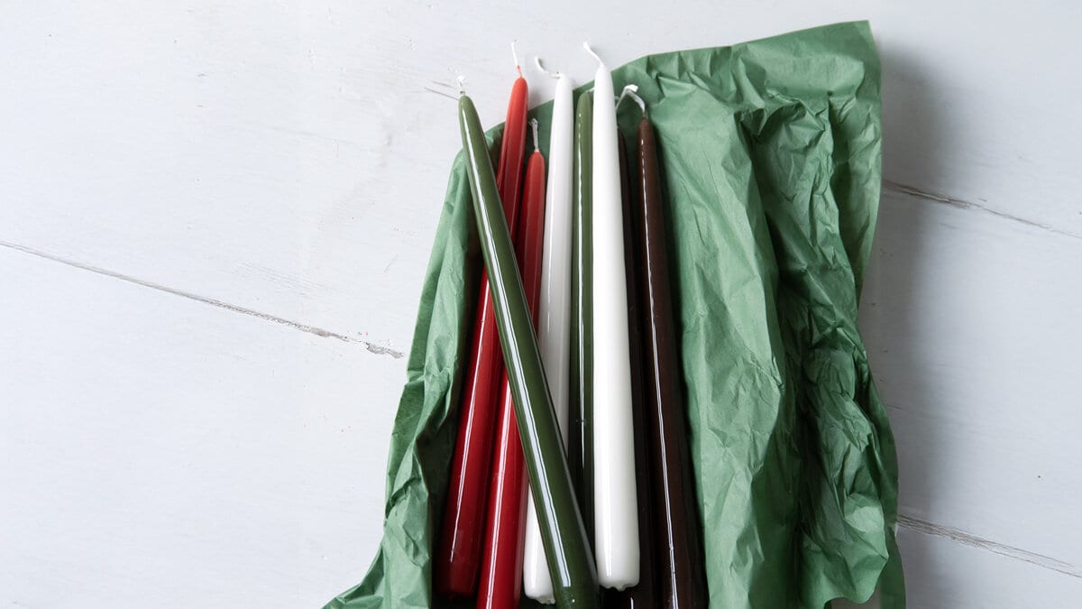 Several taper candles in different colors – red, green, white, and brown – placed on green tissue paper.