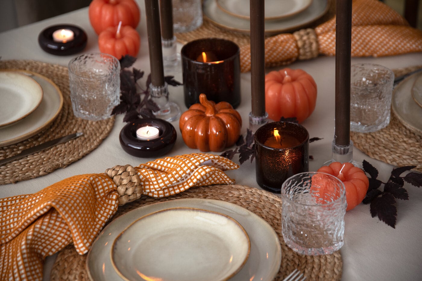 Autumn table setting with orange pumpkins, lit candles, textured glasses and an orange patterned napkin.