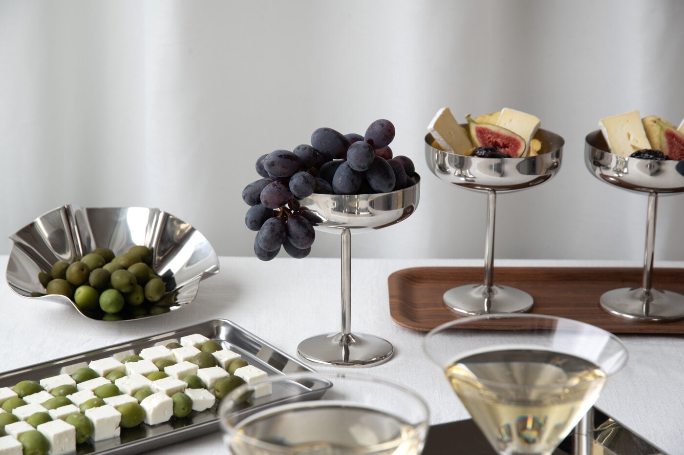 Silver-coloured serving dishes with grapes, olives, cheese and figs on a party table.