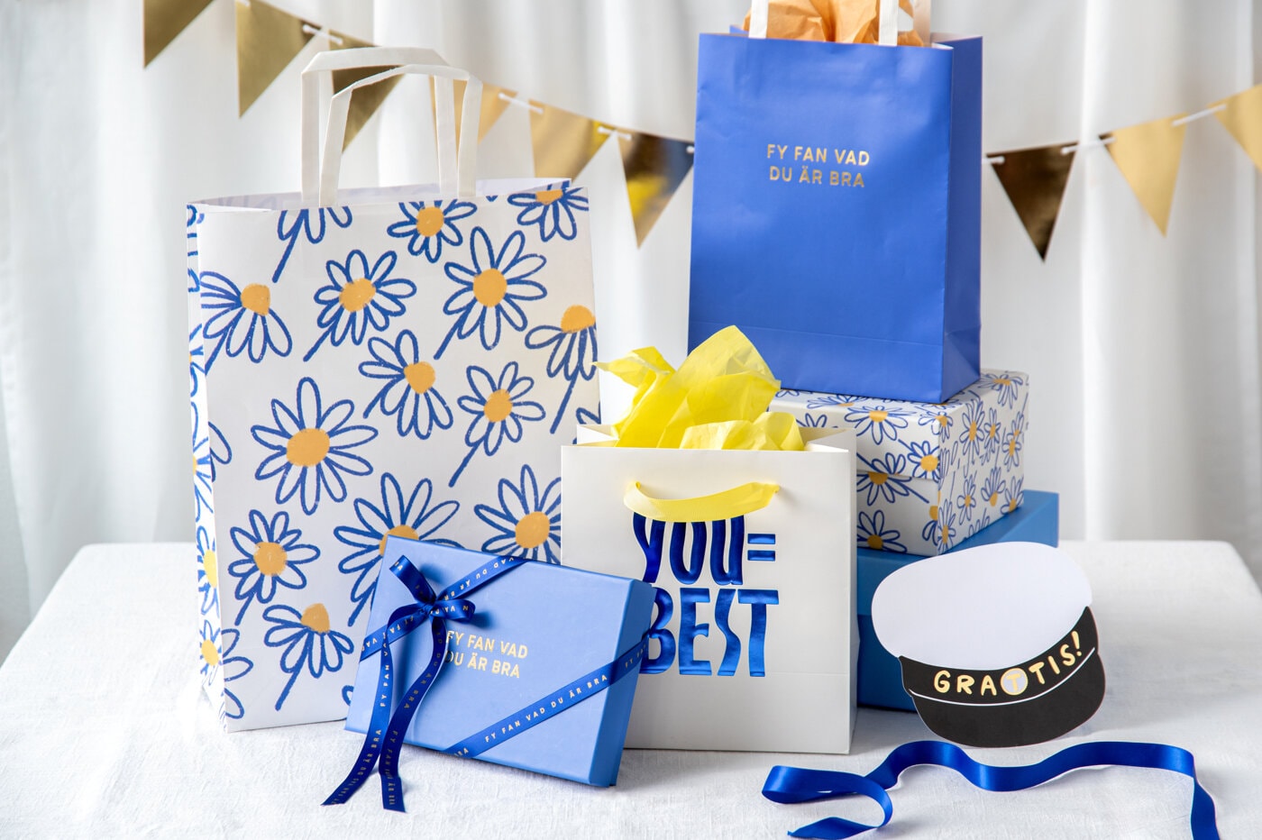 Presentpåsar och presentboxar i blått, vitt och gult med blommigt mönster uppställda tillsammans.
