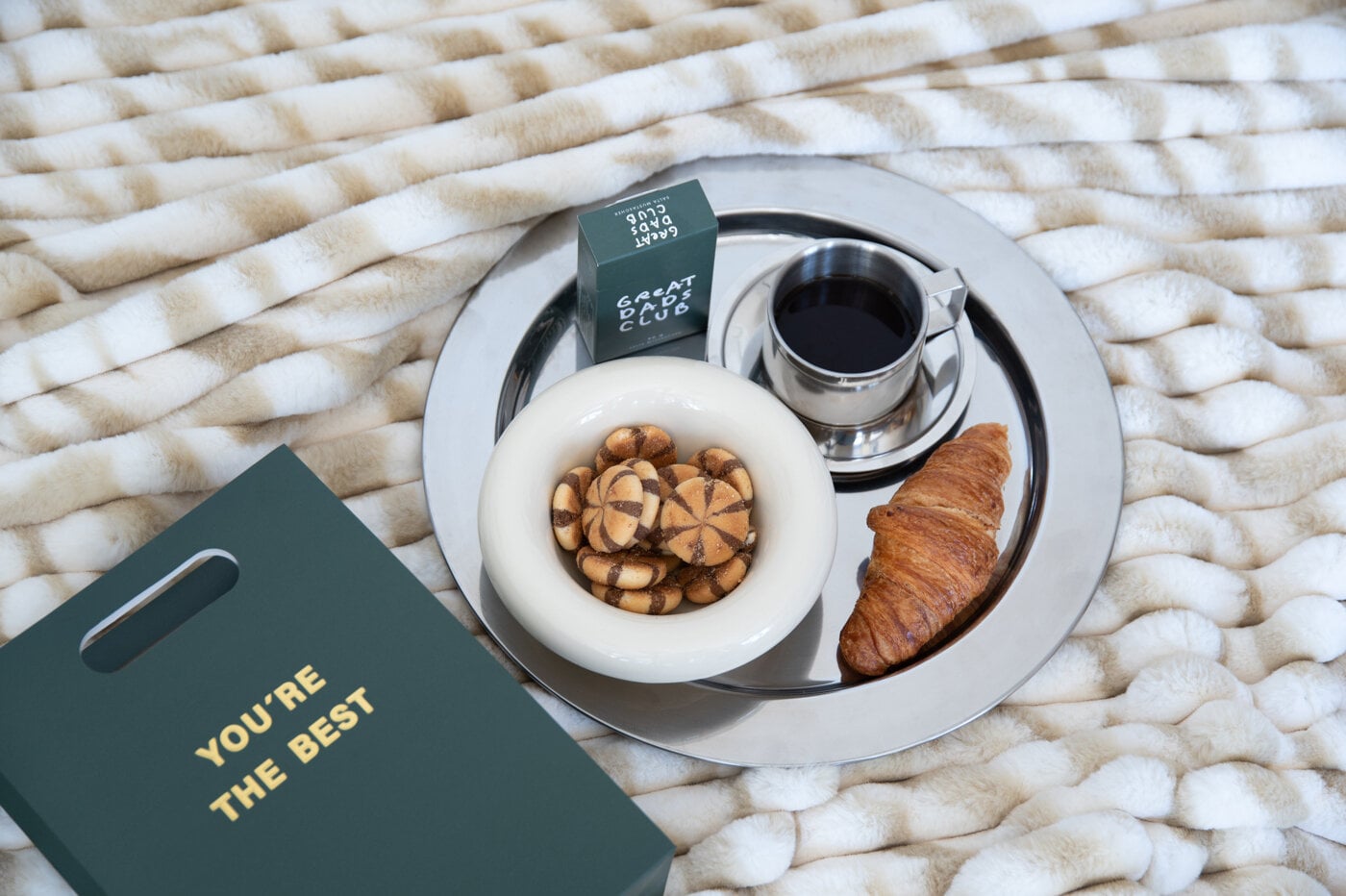 Tray with coffee, a croissant, cookies, and a gift bag on a soft blanket.