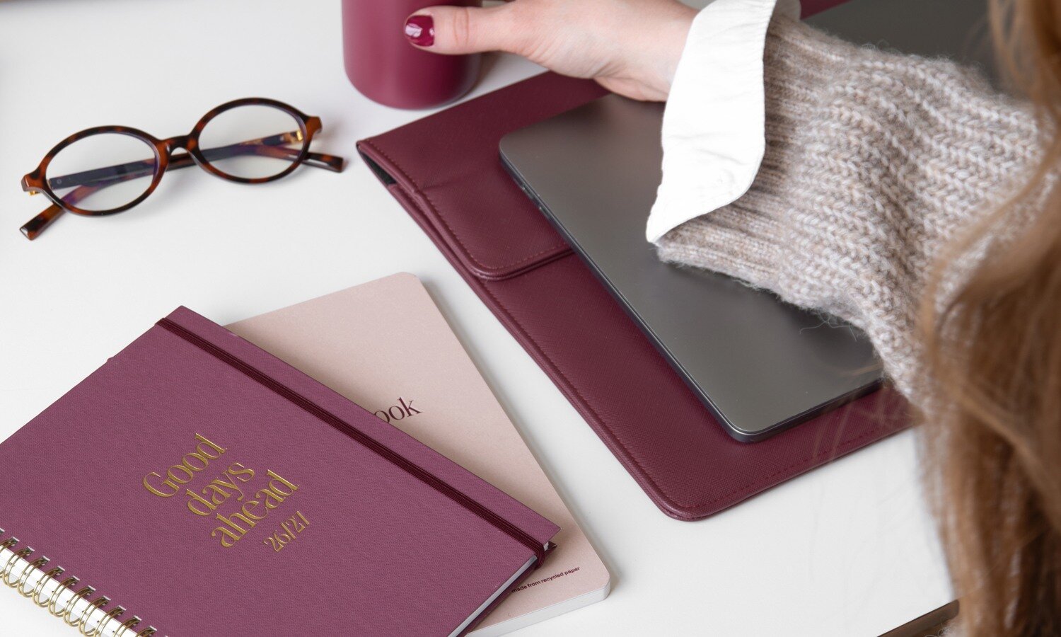 Pink and burgundy planners, notebooks, laptop and desk accessories arranged on a white surface.